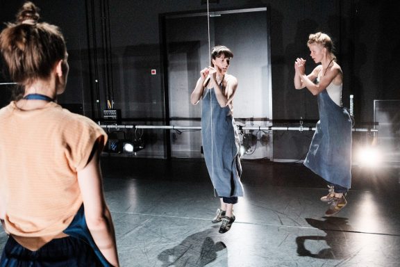 Zwei Tänzerinnen in blauen Overalls führen eine choreografierte Darbietung auf.